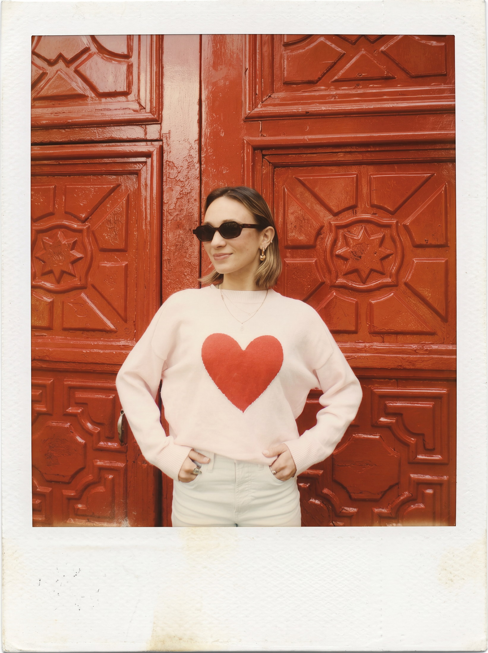 Juliet Hay in sunglasses and a heart sweater in front of a carved red door