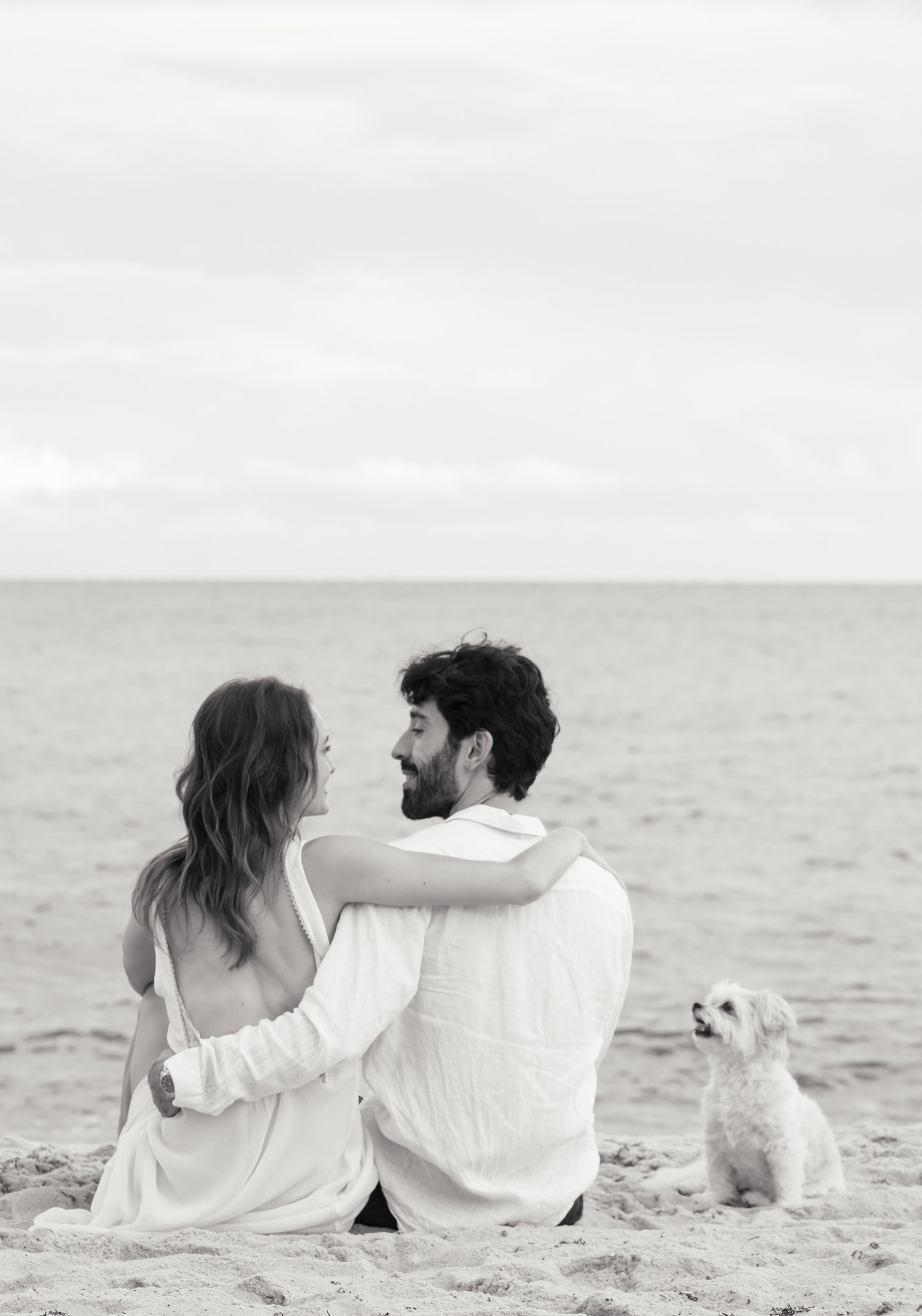 Black-and-white portrait of a couple seated with Bella beside the ocean